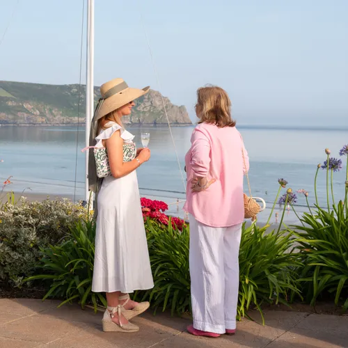 Two women in summer outfits chat beside a sailboat mast overlooking a calm bay and coastal cliffs.