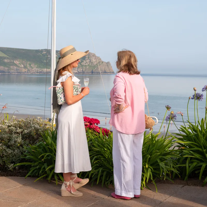 Two women in summer outfits chat beside a sailboat mast overlooking a calm bay and coastal cliffs.