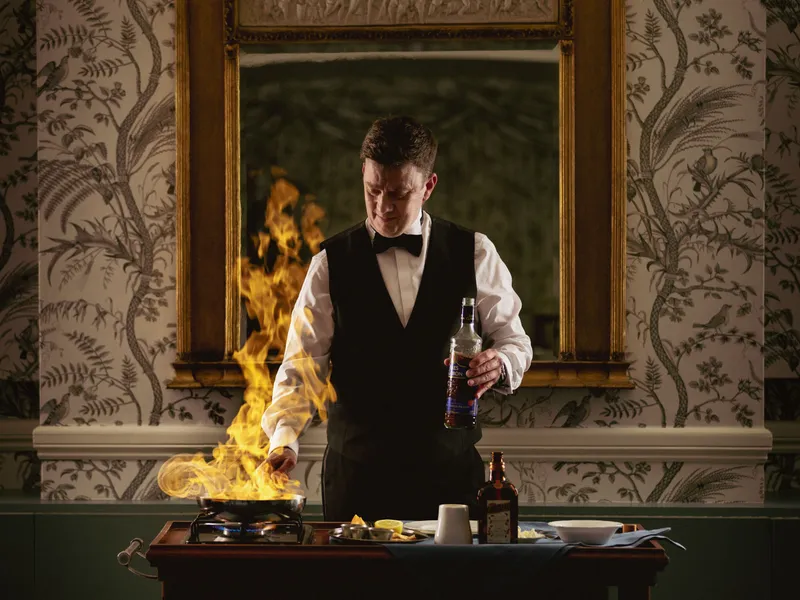 Bartender in formal attire preparing a flaming drink at a trolley while holding a bottle of whisky.