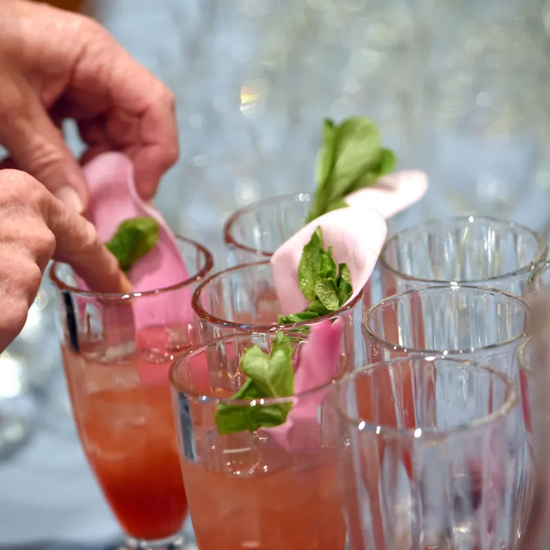 Hands garnish pink drinks in tall glasses with mint leaves and edible pink flowers.
