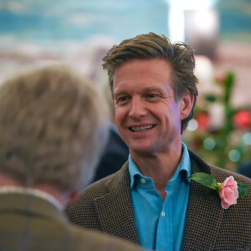 Smiling man in a blue shirt and checked jacket wearing a pink rose boutonnière at an indoor gathering