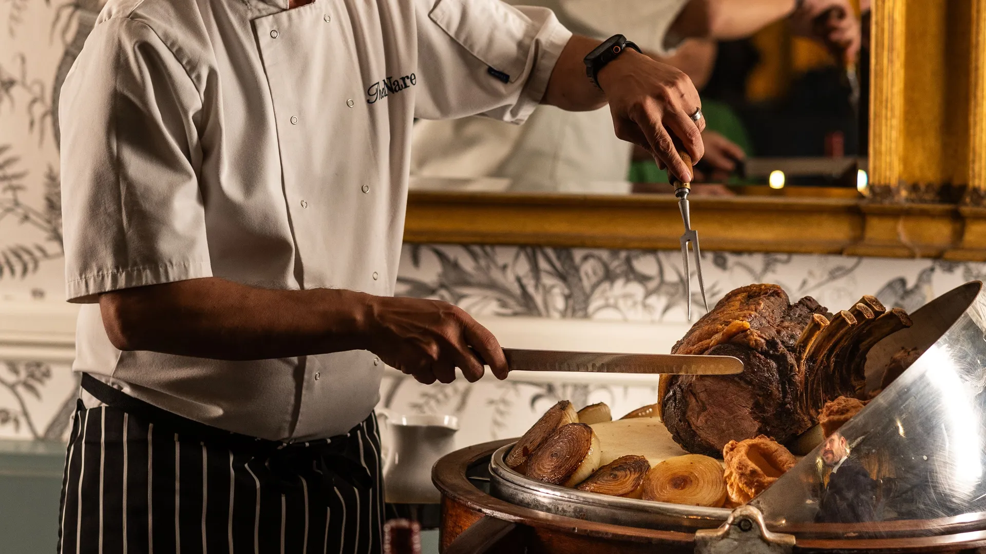 Chef carving a roast at a trolley in an elegant dining room with a large gilt mirror behind him