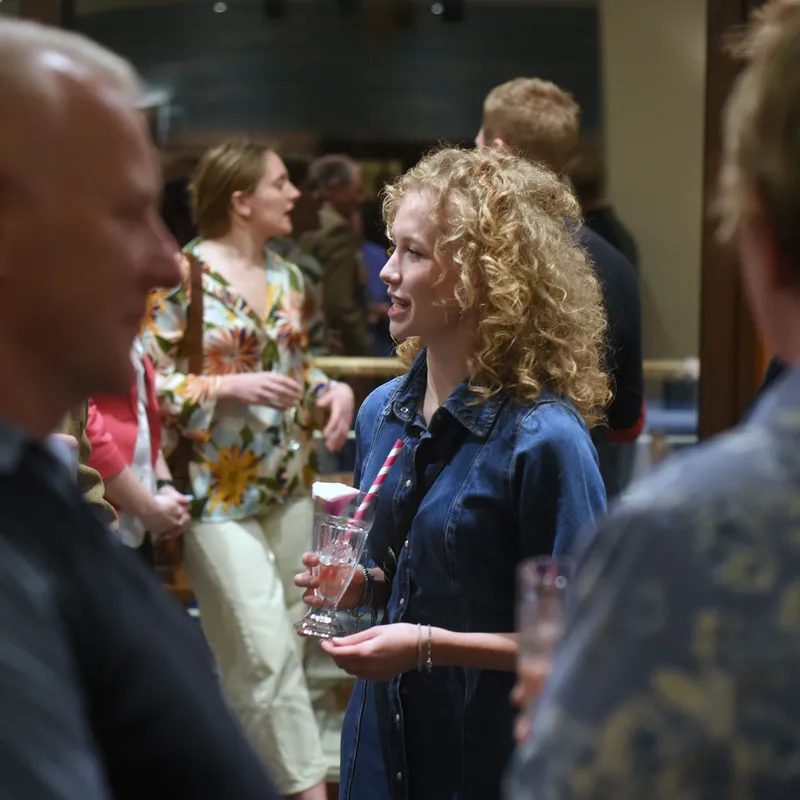 Curly-haired woman in a denim dress holding a drink and chatting at an indoor social gathering.