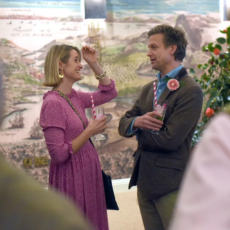 Two guests chatting with drinks at an indoor celebration beside a floral display and mural