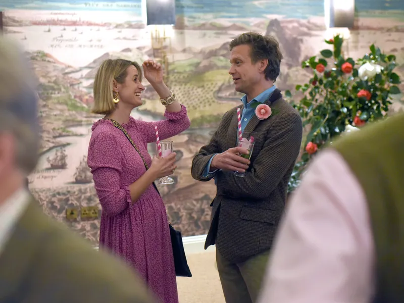 Two guests chatting with drinks at an indoor celebration beside a floral display and mural