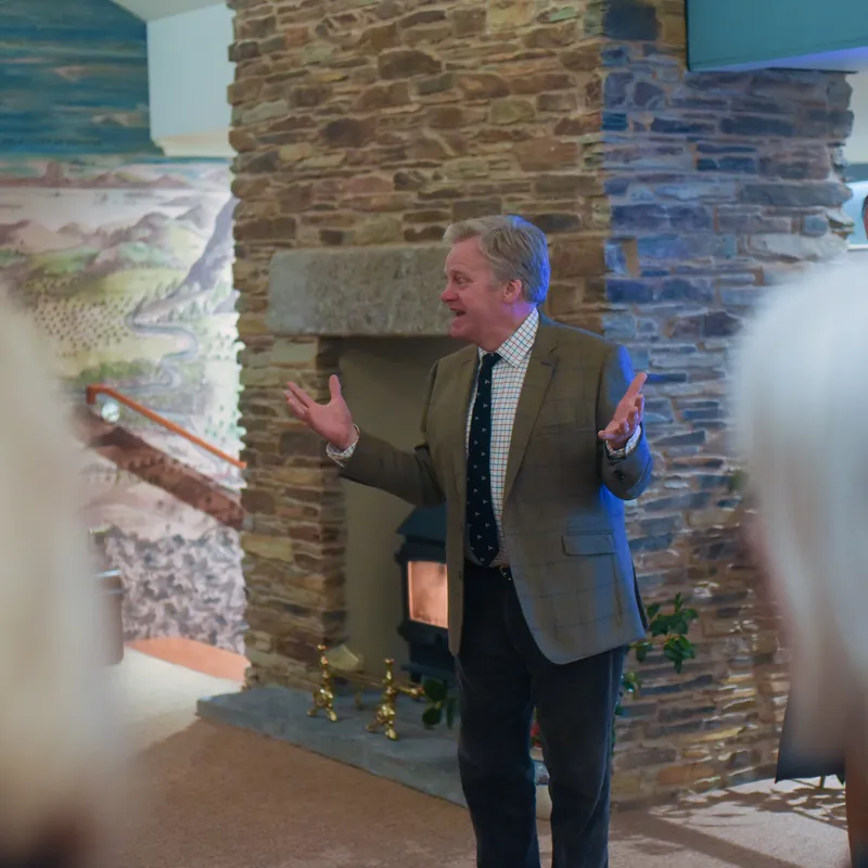 Man speaking with open hands beside a stone fireplace in a rustic indoor venue