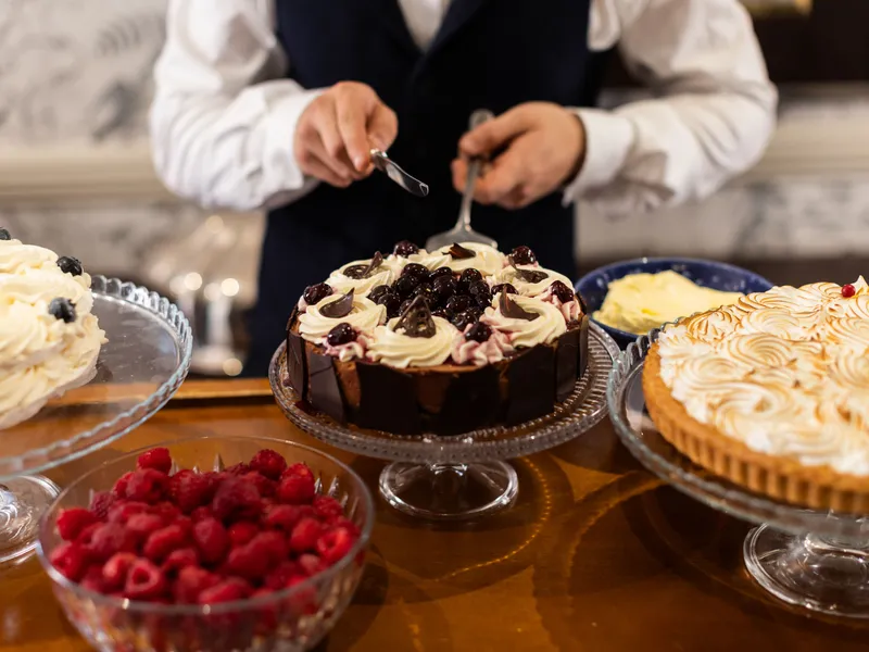 A server slices a decorated chocolate cake surrounded by fruit tarts, cream cakes and fresh raspberries on a dessert table.