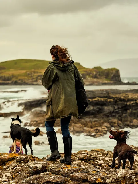 Person in a green coat standing with two dogs on a rocky coastline beneath a cloudy sky