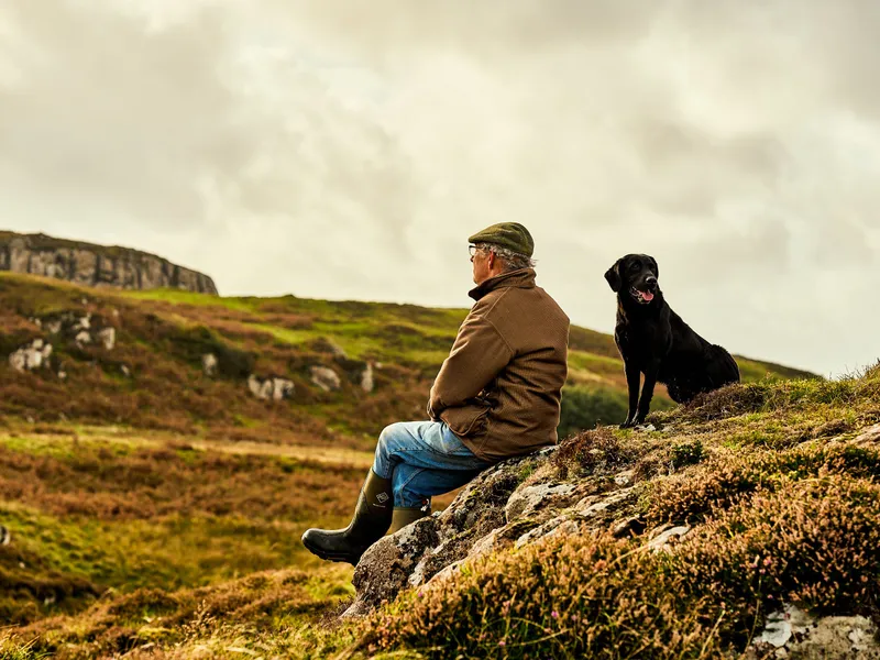 Man in a flat cap sitting on a rocky hillside beside a black Labrador, overlooking moorland beneath a cloudy sky.