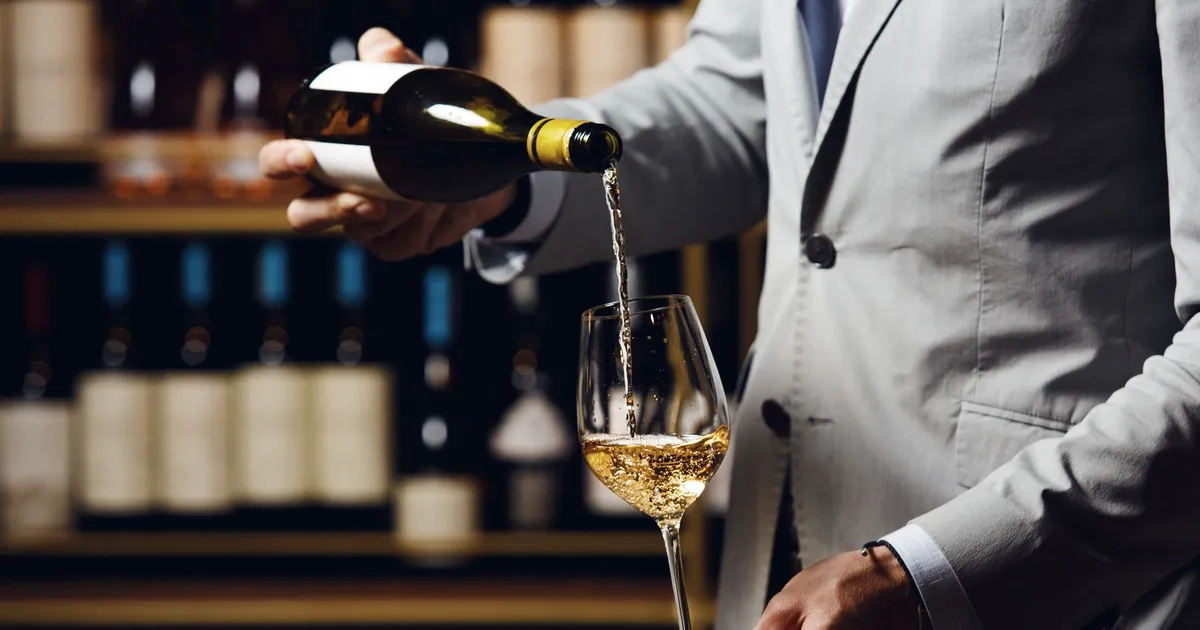 Sommelier in a grey suit pouring white wine from a bottle into a stemmed glass in a wine shop.