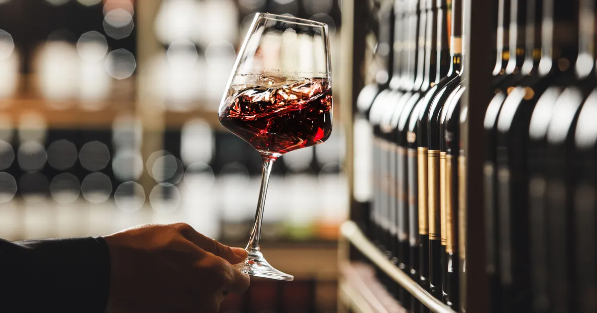 Hand holding a glass of red wine beside shelves of wine bottles in a softly lit wine shop.