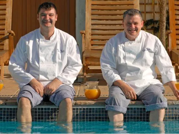 Two smiling chefs in white jackets sit with their feet in a swimming pool beside glasses of orange juice.