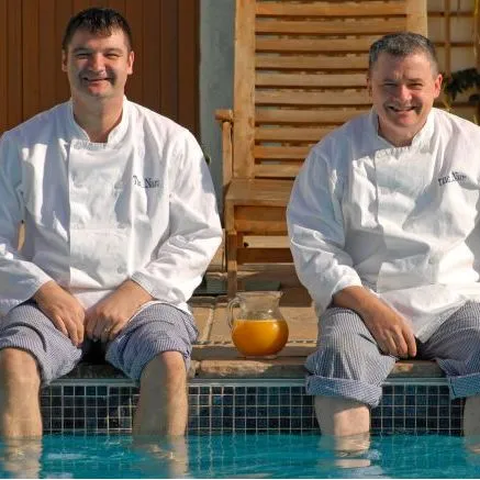 Two smiling chefs in white jackets sit with their feet in a swimming pool beside glasses of orange juice.