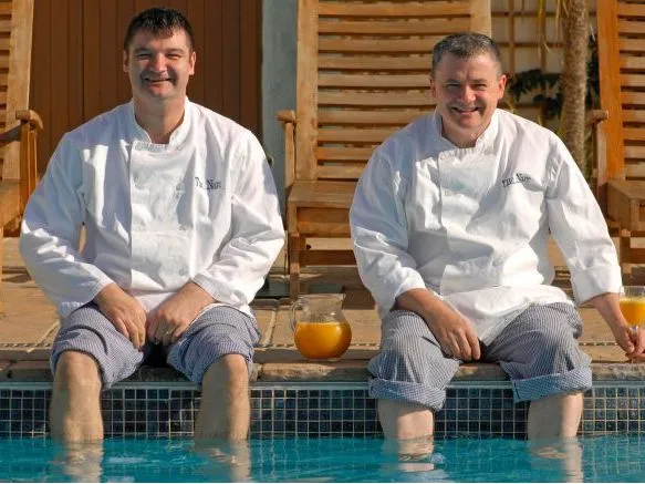 Two smiling chefs in white jackets sit with their feet in a swimming pool beside glasses of orange juice.