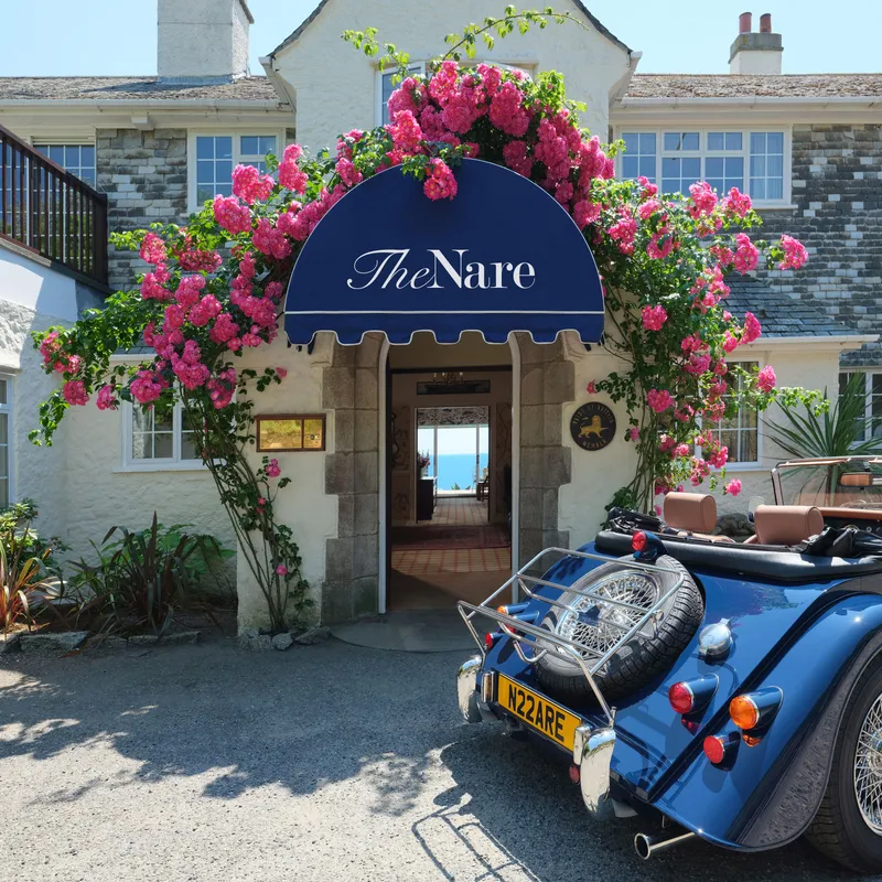 Entrance to The Nare hotel framed by bright pink climbing roses, with a blue vintage convertible parked outside.