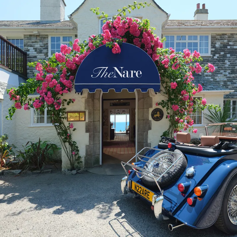 Entrance to The Nare hotel framed by bright pink climbing roses, with a blue vintage convertible parked outside.