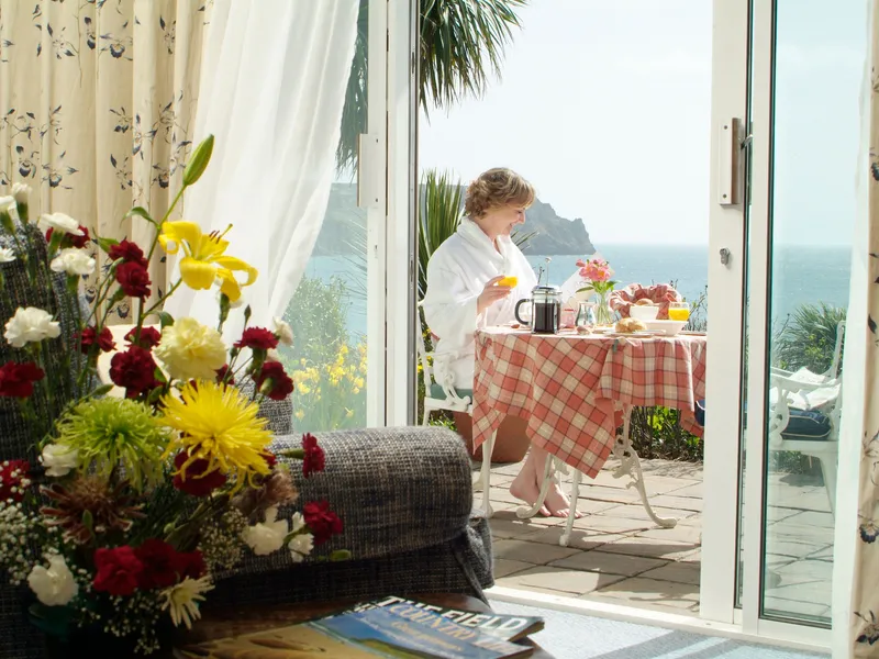 Woman in a white robe having breakfast on a seaside patio viewed through open glass doors from a flower-filled bedroom.