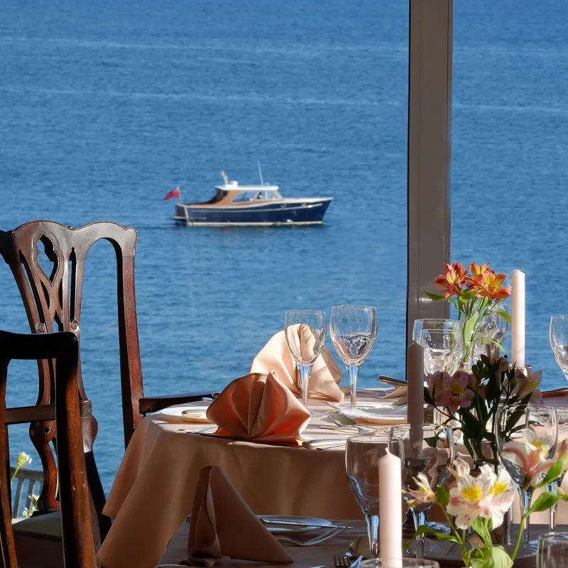 Elegant restaurant dining tables set beside large windows overlooking a blue sea and a small boat.