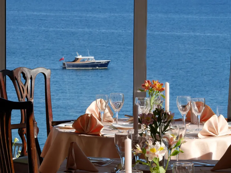 Elegant restaurant dining tables set beside large windows overlooking a blue sea and a small boat.