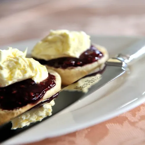 Two scones topped with clotted cream and dark berry jam on a white plate with a butter knife.