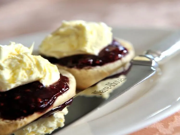 Two scones topped with clotted cream and dark berry jam on a white plate with a butter knife.