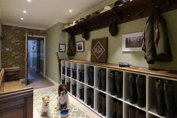 Two dogs sitting in a traditional boot room with shelves of Wellington boots, coats and hats.