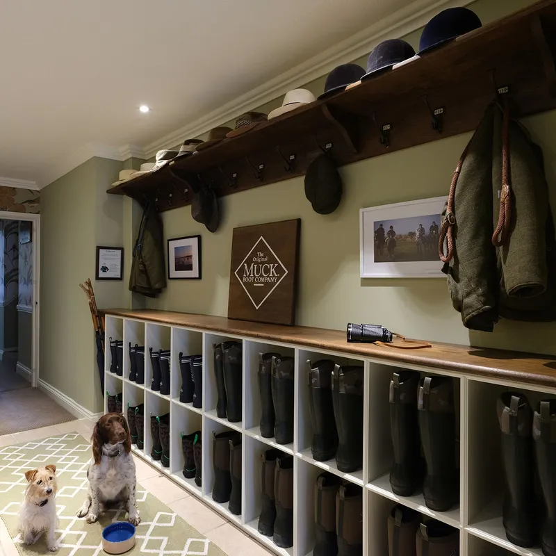 Two dogs sitting in a traditional boot room with shelves of Wellington boots, coats and hats.