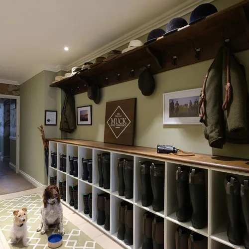 Two dogs sitting in a traditional boot room with shelves of Wellington boots, coats and hats.
