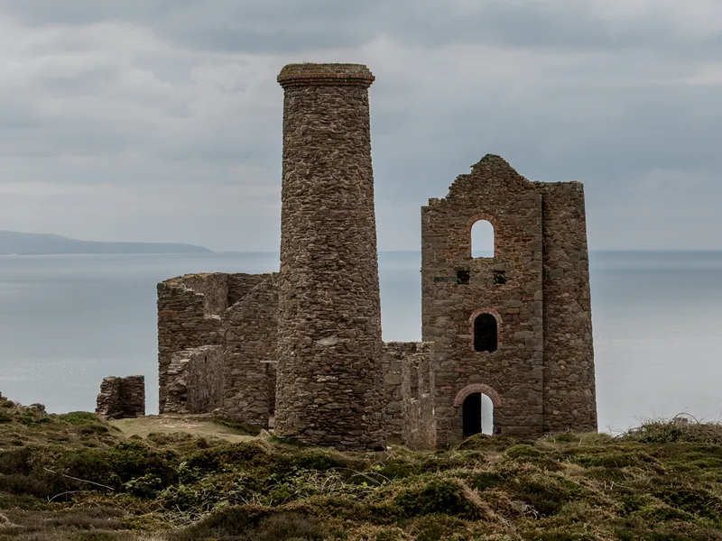 Ruins of an old stone mine engine house and chimney on a coastal hillside under a cloudy sky