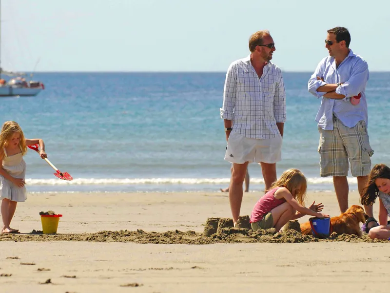 Children building sandcastles with two men nearby on Carne Beach, with a sailboat on the sea behind them.