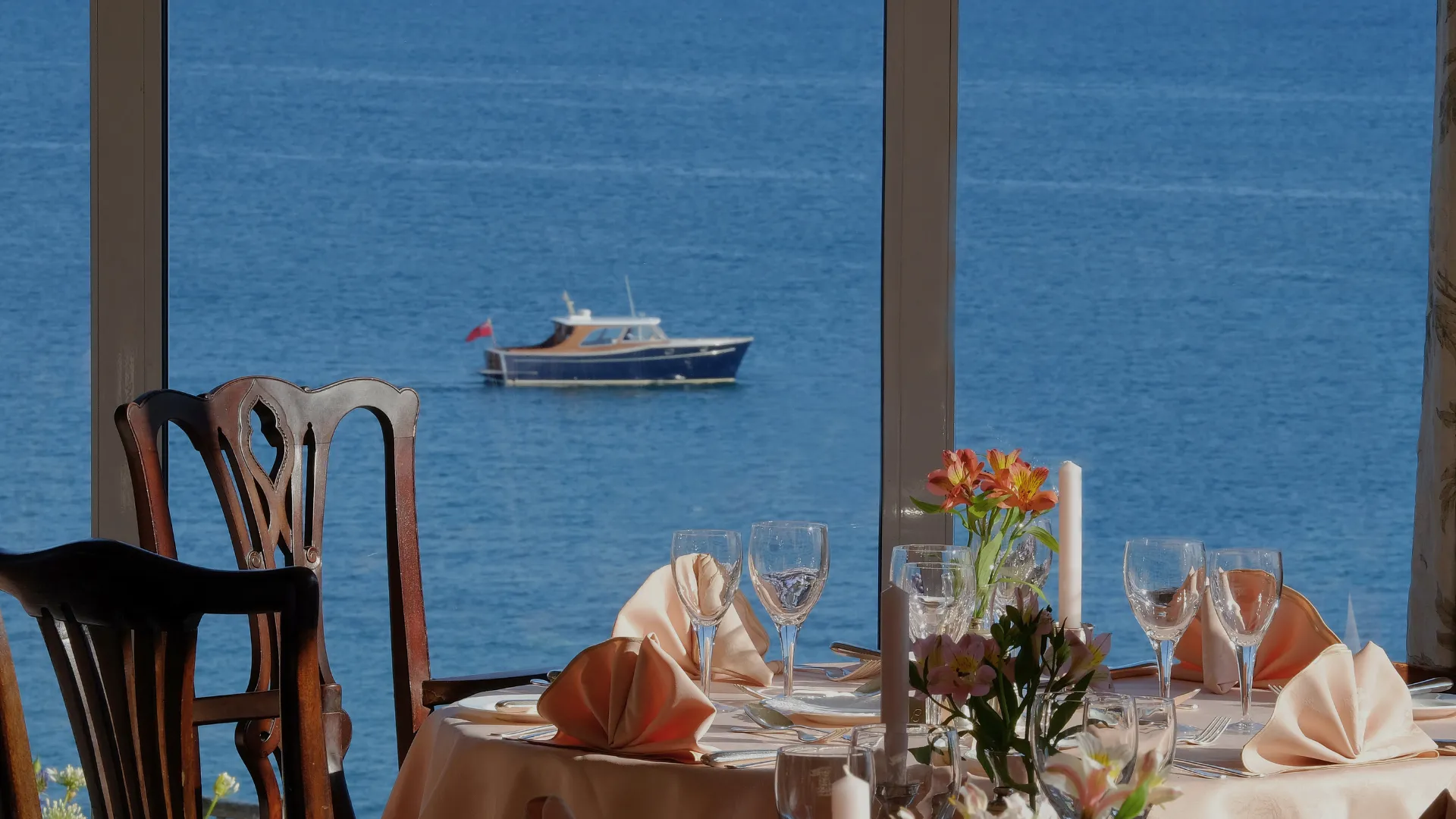Elegantly set dining table beside large windows overlooking a blue sea and passing motorboat.