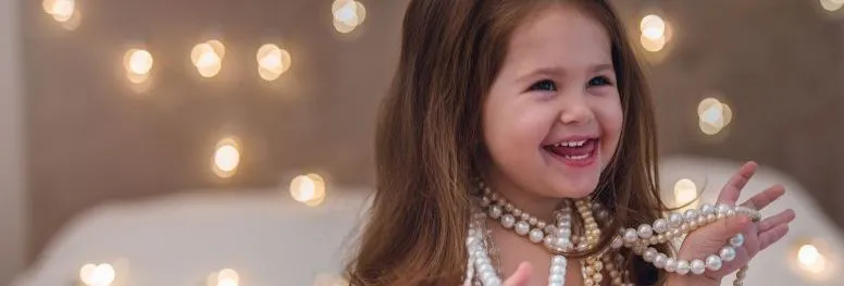 Smiling young girl wearing and holding pearl necklaces beside a bed with warm fairy lights.