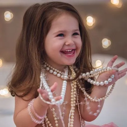 Smiling young girl wearing and holding pearl necklaces beside a bed with warm fairy lights.