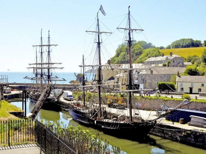 Historic tall ships moored in a Cornwall harbour beside stone cottages and green hills.