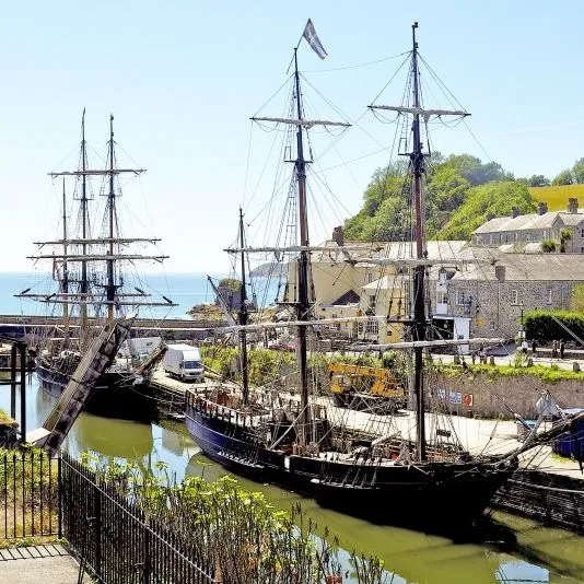 Historic tall ships moored in a Cornwall harbour beside stone cottages and green hills.
