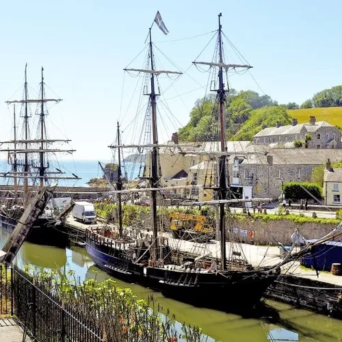 Historic tall ships moored in a Cornwall harbour beside stone cottages and green hills.