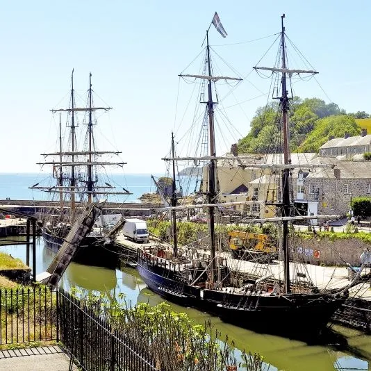 Historic tall ships moored in a Cornwall harbour beside stone cottages and green hills.