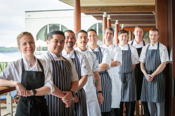 A team of chefs in white jackets and striped aprons standing together on a covered waterfront terrace.