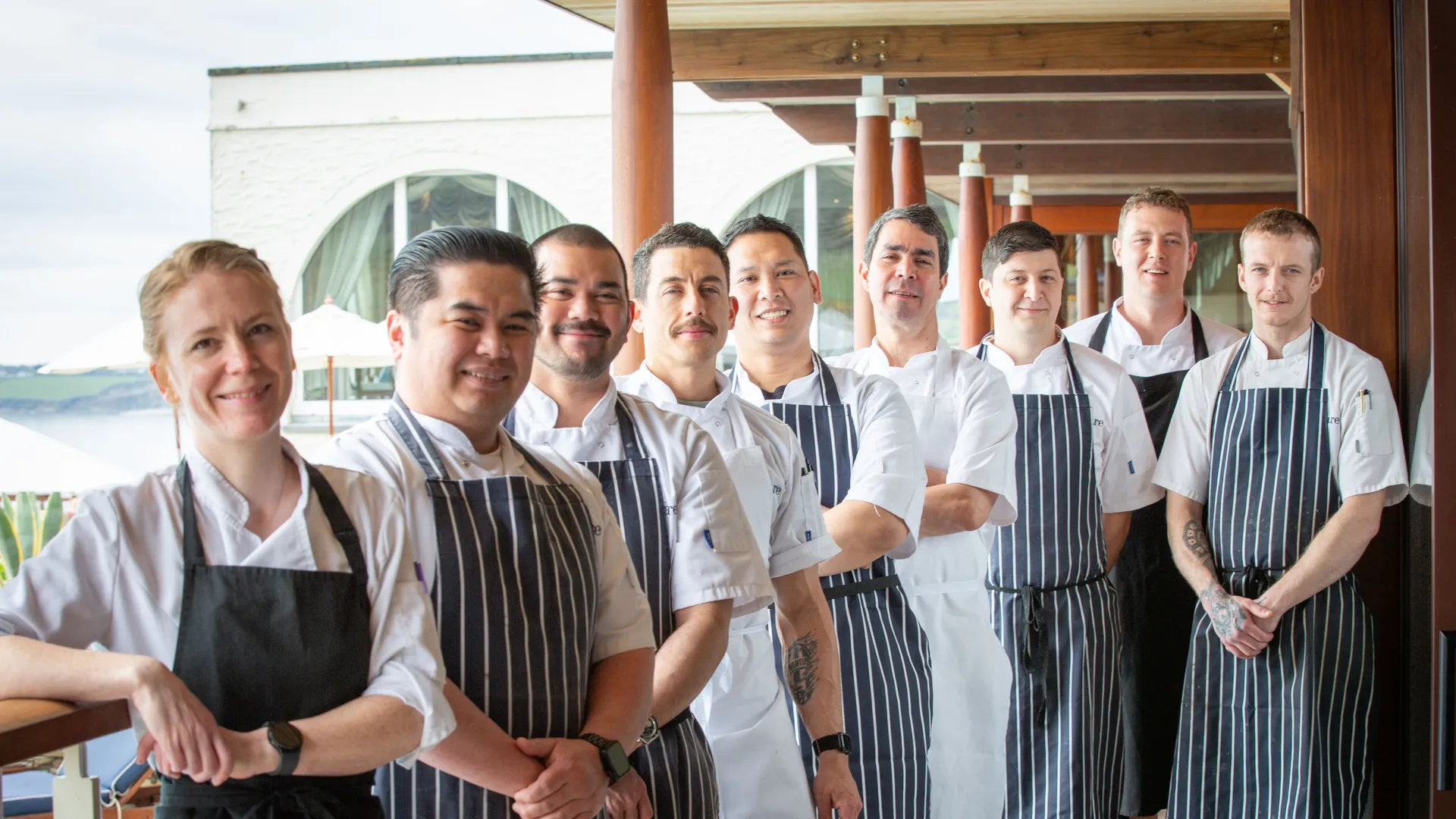 A team of chefs in white jackets and striped aprons standing together on a covered waterfront terrace.