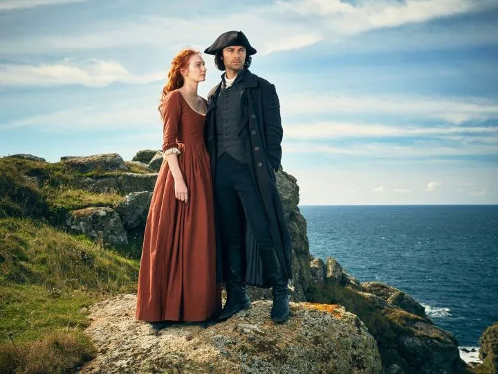A woman in a rust-coloured historical dress stands beside a man in a dark tricorn hat and coat on rocky sea cliffs.
