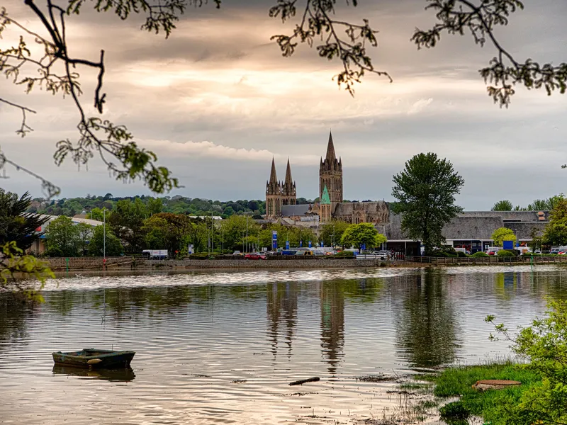Gothic cathedral reflected in a calm river beneath a cloudy sky, framed by leafy branches and a small boat.