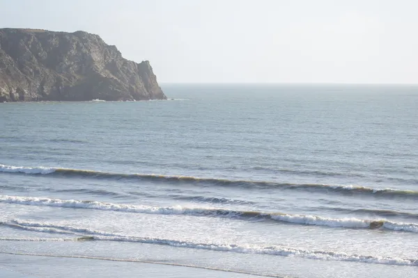 A person walking a dog on a wide sandy beach beneath a rocky headland, with gentle waves rolling in.