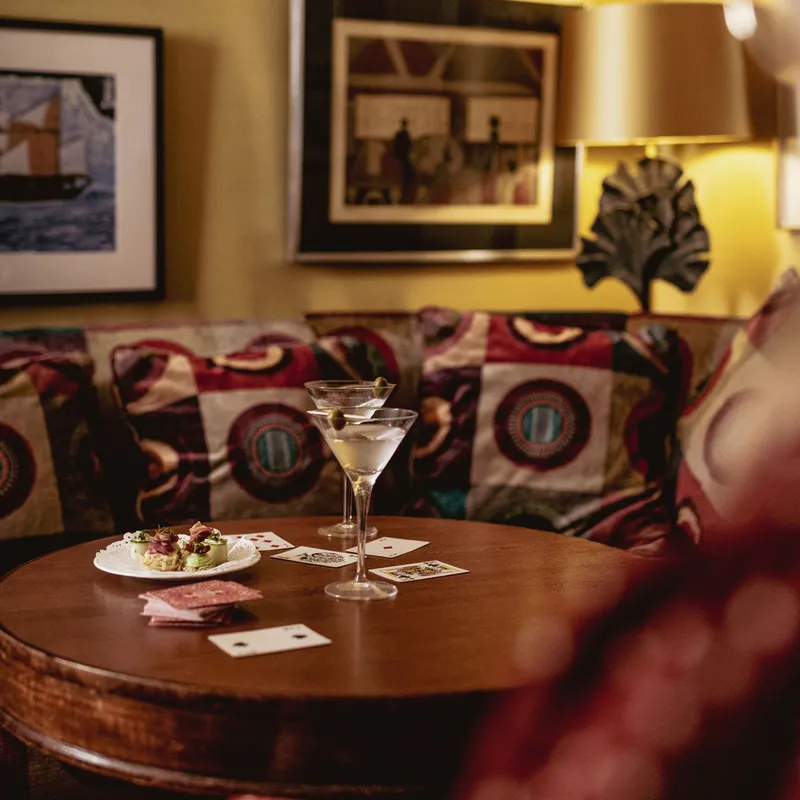 Two martini glasses, playing cards and a plate of canapés on a round wooden table beside a patterned sofa.
