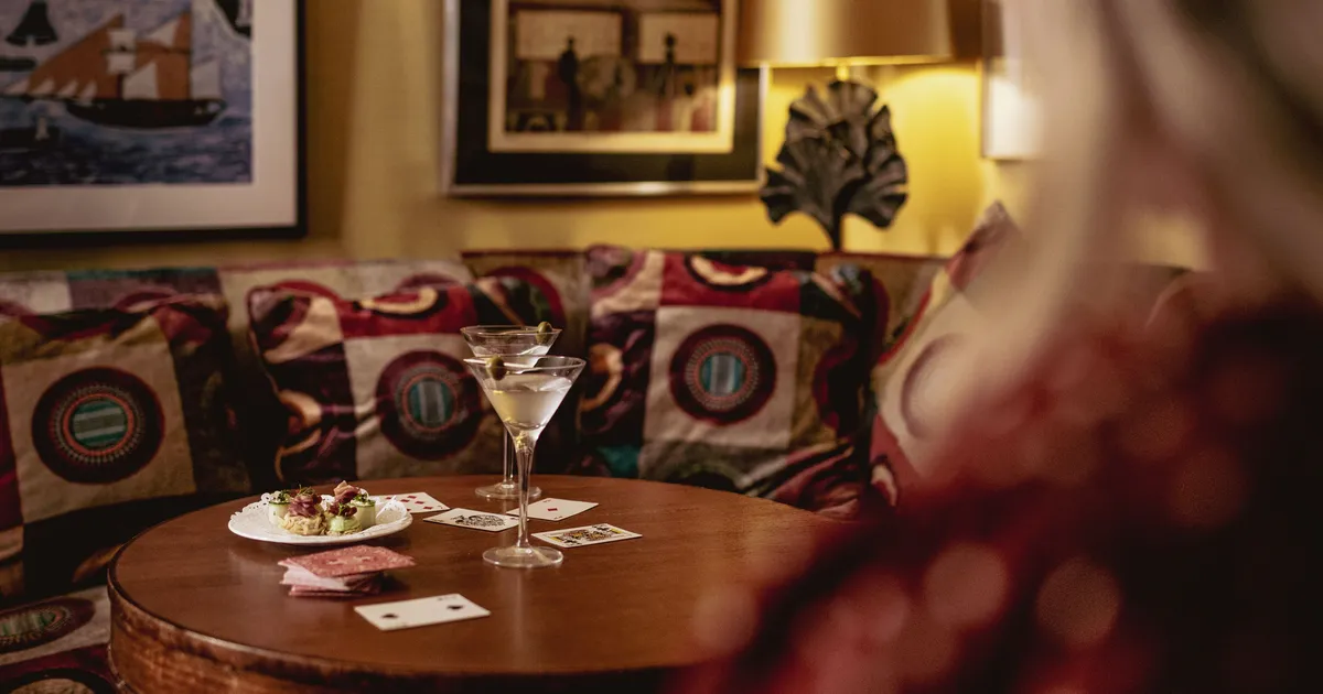 Two martini glasses, playing cards and a plate of canapés on a round wooden table beside a patterned sofa.