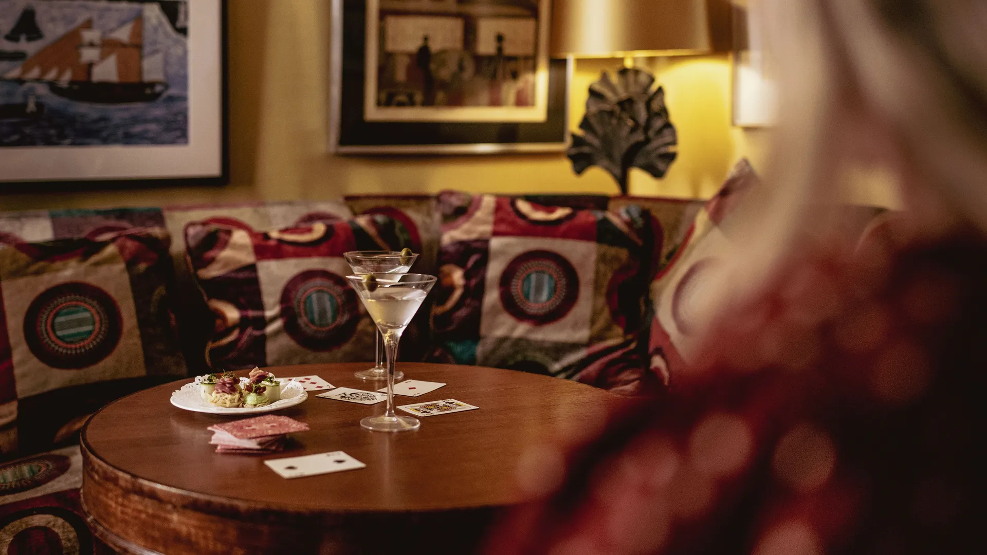 Two martini glasses, playing cards and a plate of canapés on a round wooden table beside a patterned sofa.