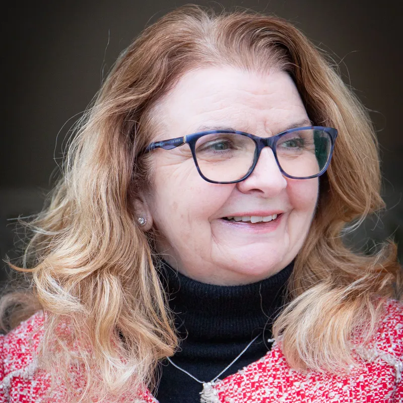 Smiling woman with long light brown hair and blue glasses, wearing a red and white jacket over a black top.