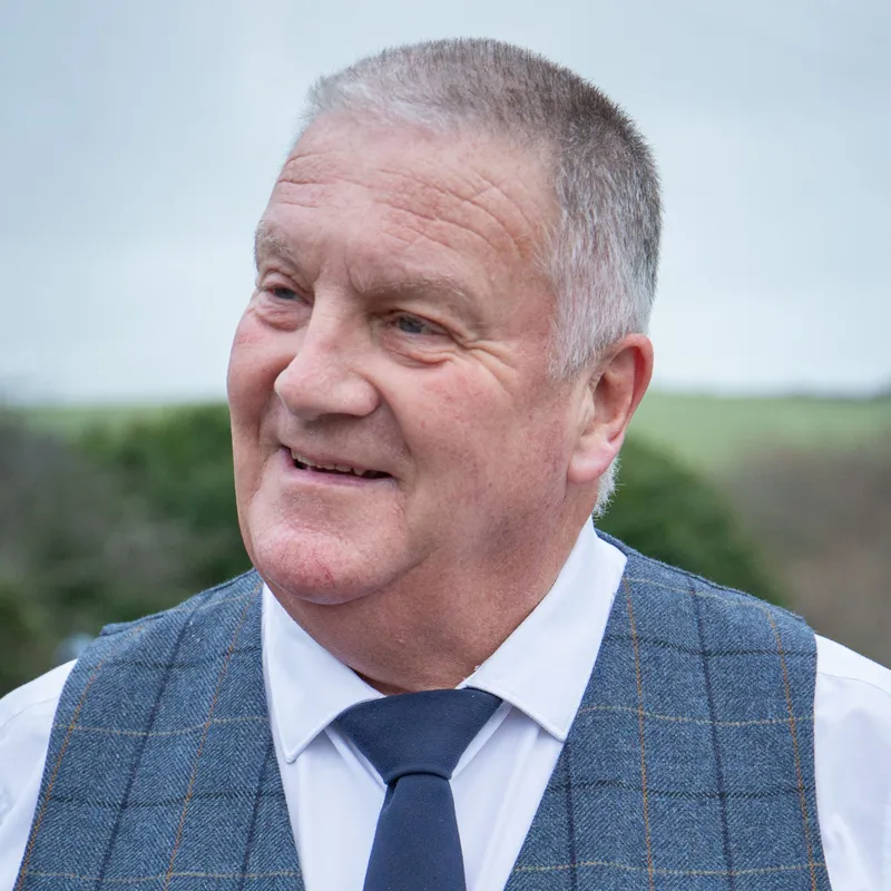 Close-up portrait of a smiling man in a blue checked waistcoat, white shirt and navy tie outdoors.