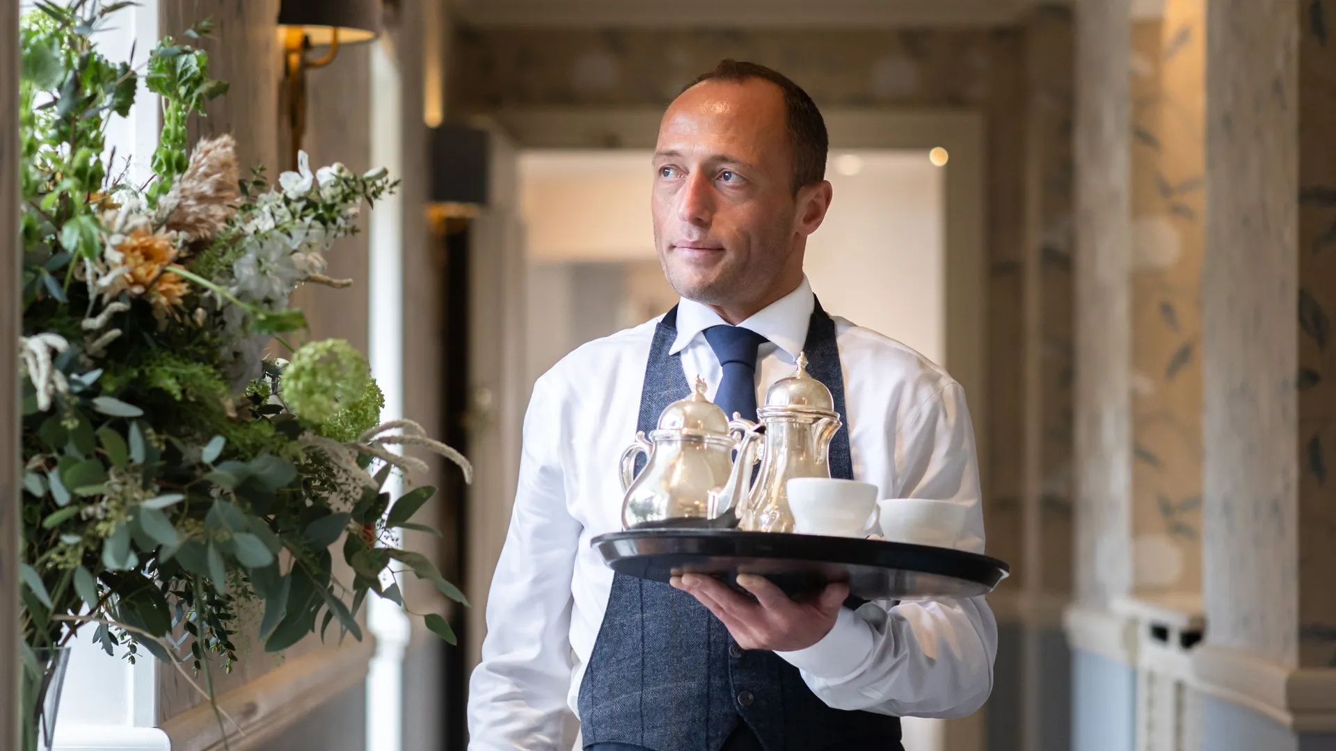 Waiter in a navy apron carrying a tray with silver teapots and white cups through an elegant hallway