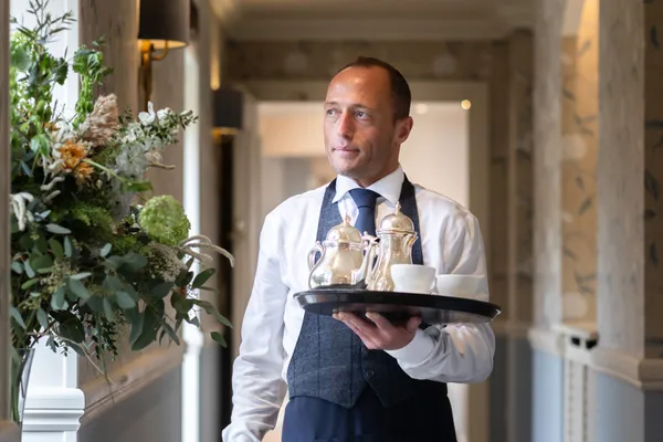 Waiter in a navy apron carrying a tray with silver teapots and white cups through an elegant hallway