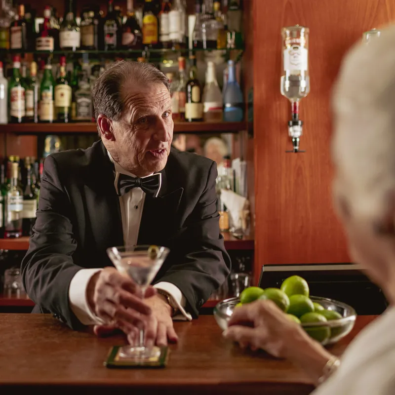 Bartender in a black dinner jacket serves a martini to a guest at a wood-panelled bar.